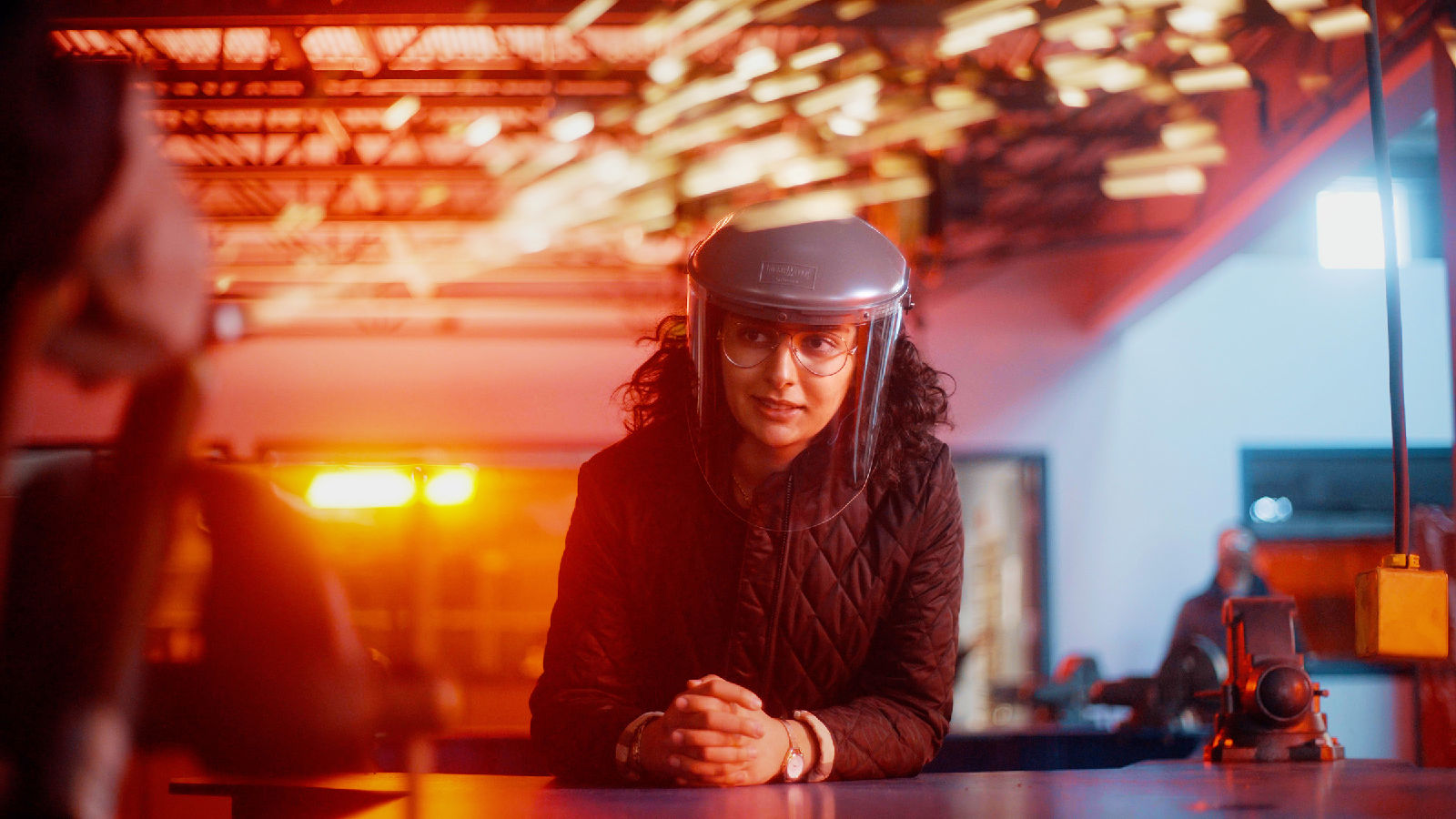 A woman with a face shield looks on as sparks are generated in a factory setting.